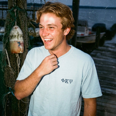 Smiling young man on a dock wearing a light blue t-shirt with small Greek letters printed on the left chest.