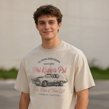 Young man wearing a vintage-style beige graphic T-shirt featuring a retro muscle car illustration and cursive text, standing outdoors with a neutral blurred background.