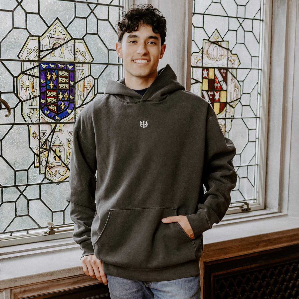 College-aged man wearing the Phi Kappa Psi embroidered black hoodie, standing in front of stained glass windows with fraternity-style crest designs.