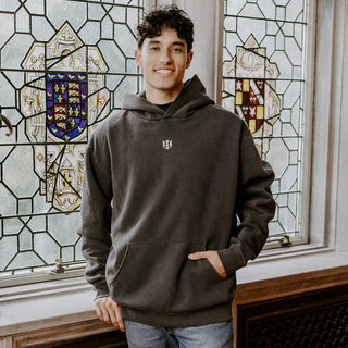 College-aged man wearing the Phi Kappa Psi embroidered black hoodie, standing in front of stained glass windows with fraternity-style crest designs.