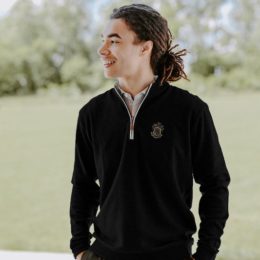 College-aged man smiles and looks off to the side while wearing a black quarter-zip pullover embroidered with a fraternity crest. He stands outdoors in a bright, grassy setting.