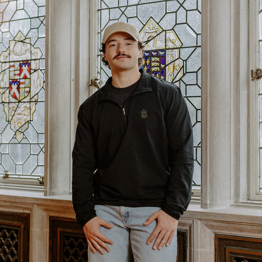 Male model wearing the black Phi Kappa Psi Embroidered Luxe Quarter Zip in front of stained glass windows with fraternity crests.