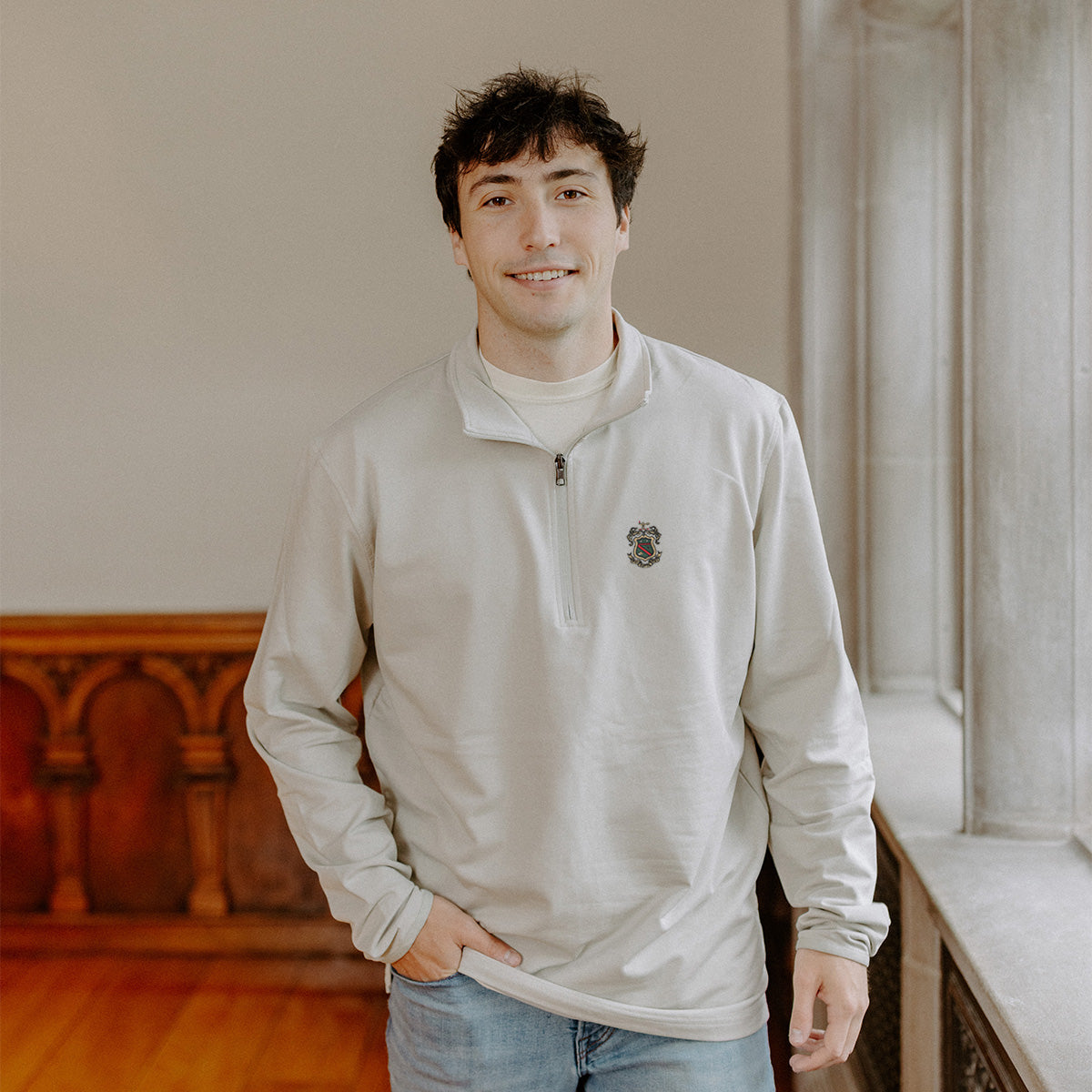 Male model wearing the smoke-colored Phi Kappa Psi Embroidered Luxe Quarter Zip with crest embroidery, standing by a window in a formal setting.