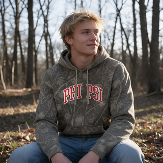 College-aged male wearing the fraternity Deadwood Camo Hoodie with red and white Phi Kappa Psi lettering, seated outdoors in a wooded setting.