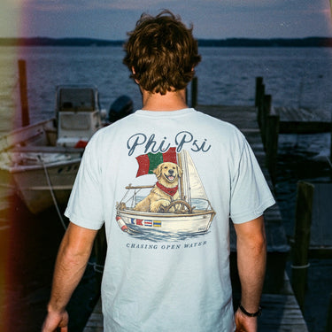 Young man standing on a dock at sunset wearing a light blue t-shirt featuring a golden retriever steering a sailboat with a flag and the phrase “Chasing Open Water” on the back.
