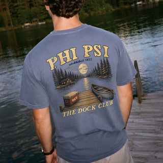Young man wearing a blue graphic tee standing on a lakeside dock, showing the back design